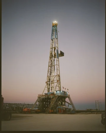 Tall oil drilling rig structure against the sky at dusk. Towering metal framework with lights on top. Industrial machinery in open setting.