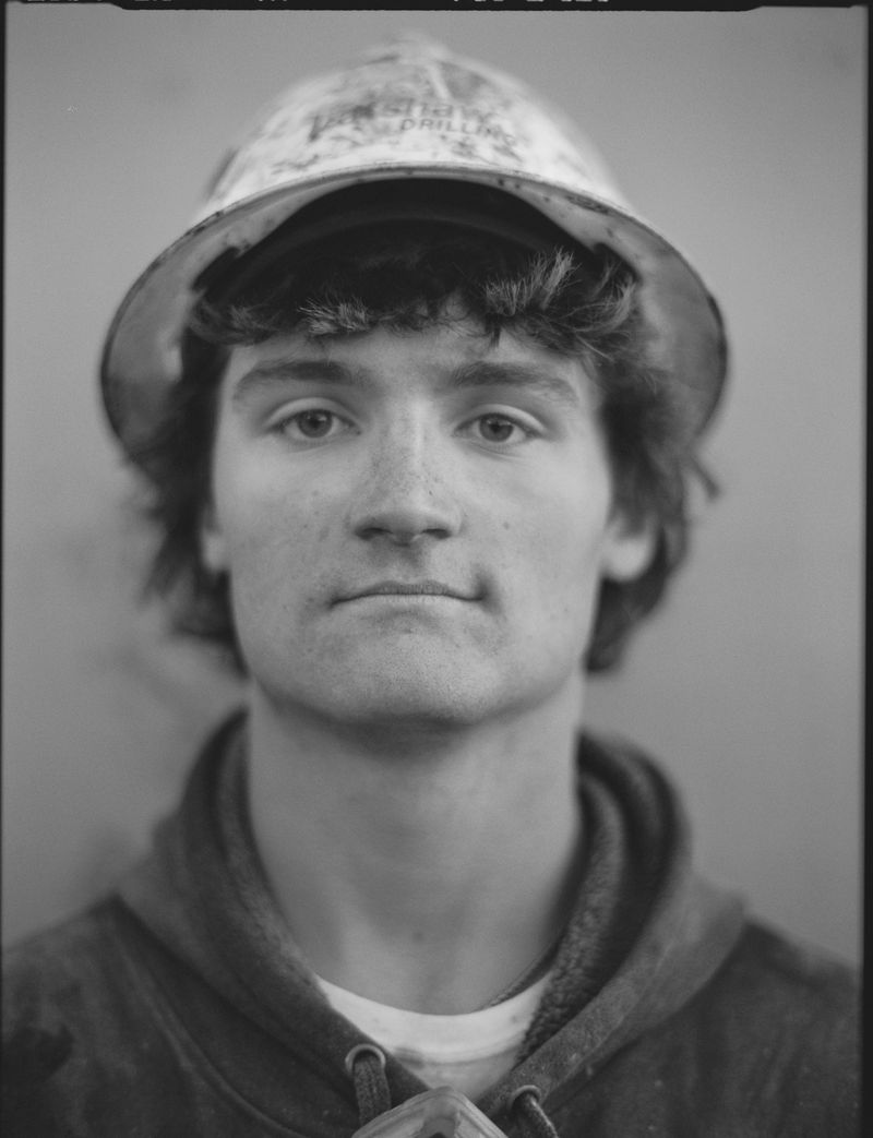 Monochromatic portrait of a young adult male wearing a hard hat and hoodie, captured in a close-up shot.