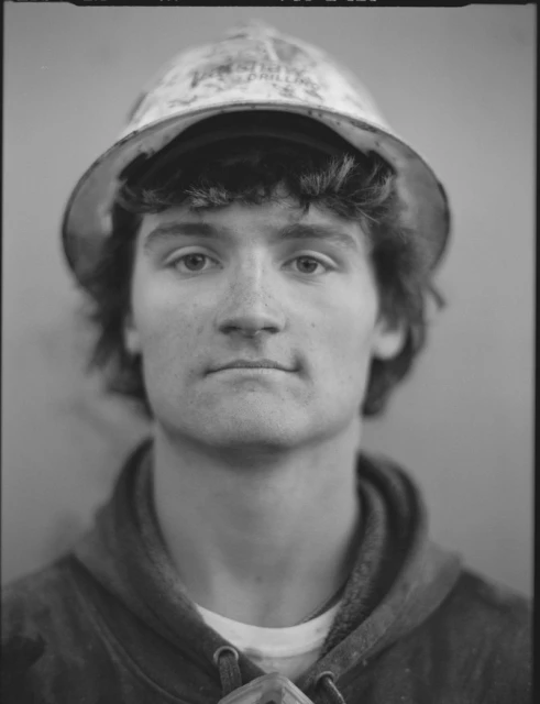 Monochromatic portrait of a young adult male wearing a hard hat and hoodie, captured in a close-up shot.