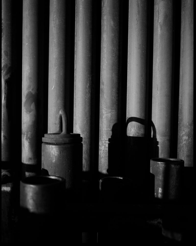 Monochrome image featuring an arrangement of industrial pipes and metal containers creating a study in texture, shadow, and form.