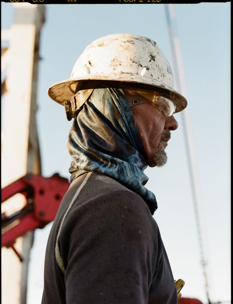 Profile portrait of a middle-aged industrial worker wearing a dirty white hard hat, safety glasses, and a scarf outdoors during the day.
