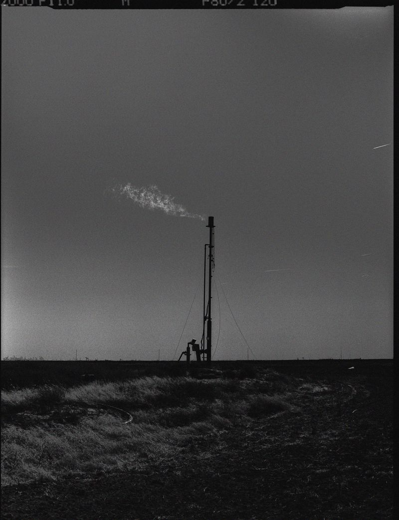 Industrial flare stack burns gas in an open field under a bright sky. The landscape is monochrome and desolate.
