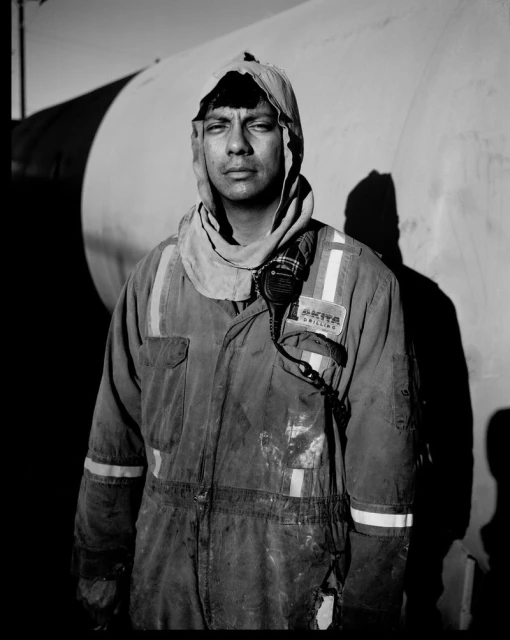 Monochrome portrait of a man in a protective suit with a serious expression, standing outdoors in front of a large, plain tank.