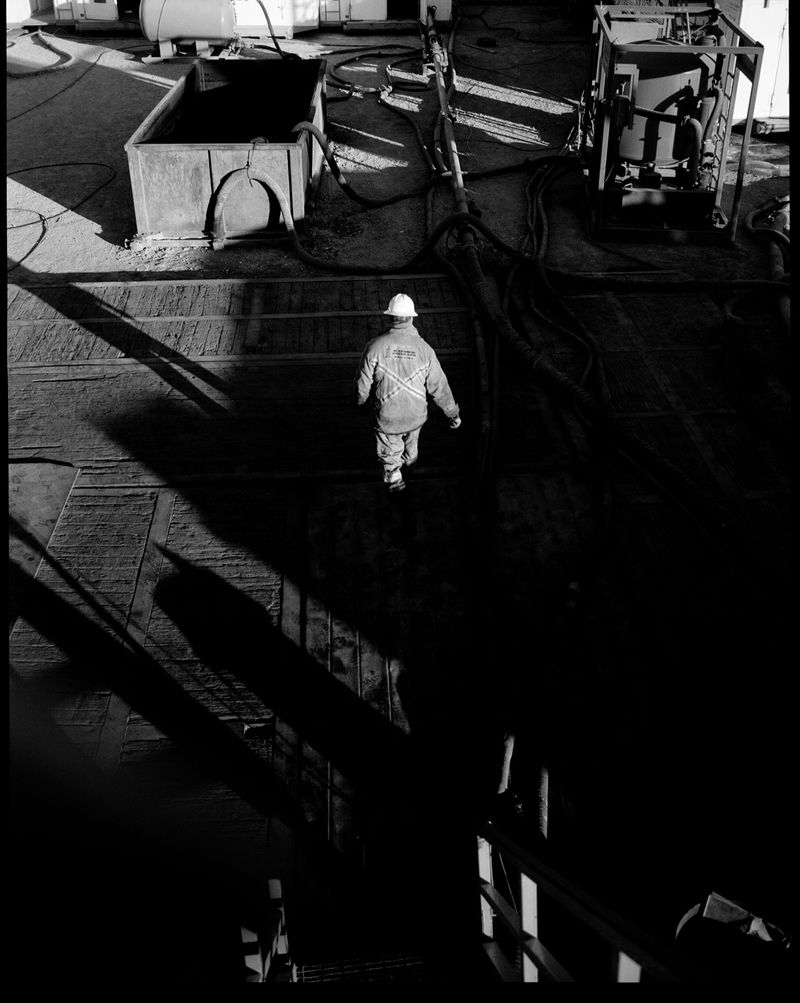 Monochromatic shot of a worker in a hard hat walking across an industrial surface surrounded by equipment, shadows, and cables.