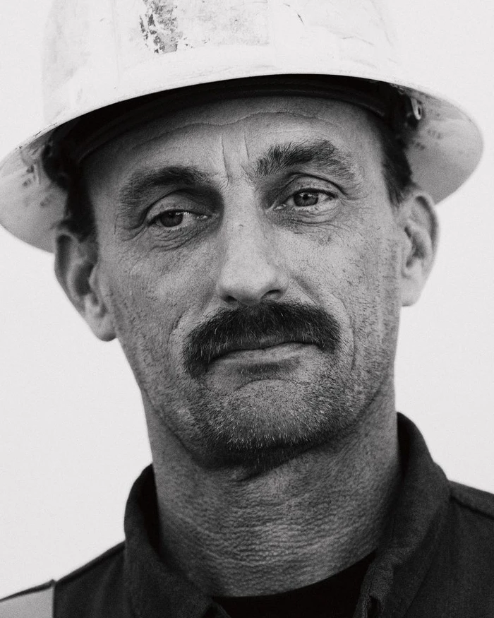 Close-up monochrome portrait of a man wearing a white hard hat and dark shirt with a mustache.