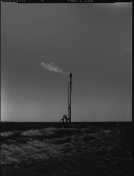 Monochrome vertical film frame showing an industrial flare stack venting a plume of smoke against an overcast sky above flat terrain.