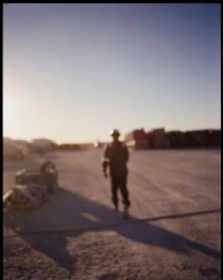 Out-of-focus figure standing on an expansive open ground at sunset with long shadows stretching across the dirt.