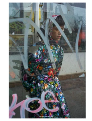 Model in floral sequin coat with matching headscarf viewed through a glass window covered in graffiti