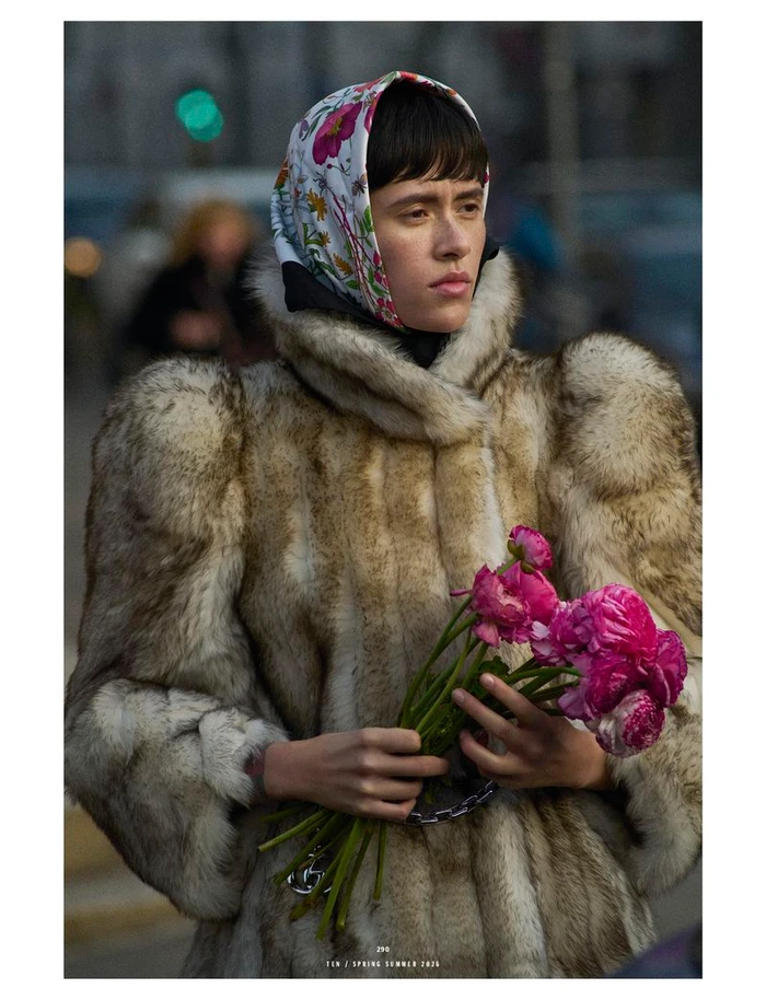 Model wearing a fur coat and floral headscarf holding a bouquet of pink flowers outdoors