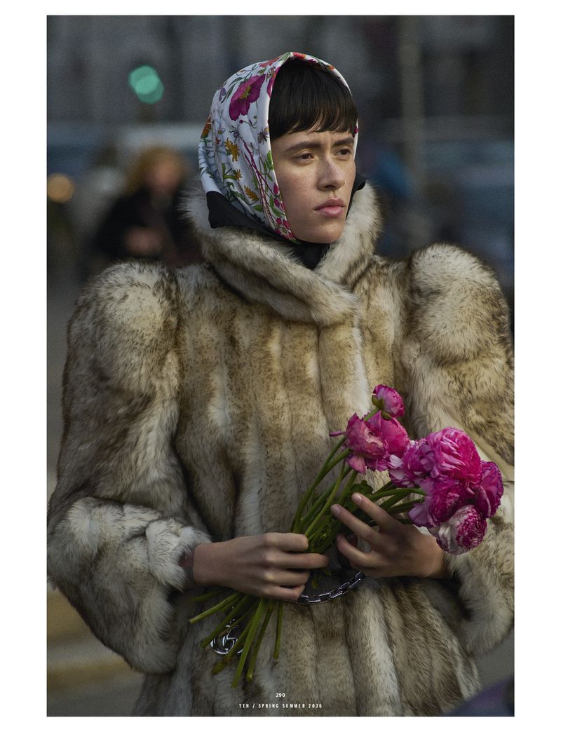 Model with short dark hair wears a printed headscarf and a textured fur coat while holding a bouquet of bright pink ranunculus flowers.