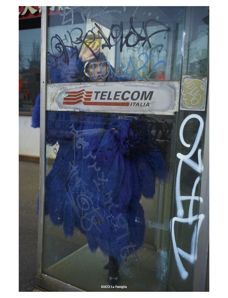Person inside graffiti-covered telephone booth with 'Telecom Italia' logo, blue furry installation, and street setting.