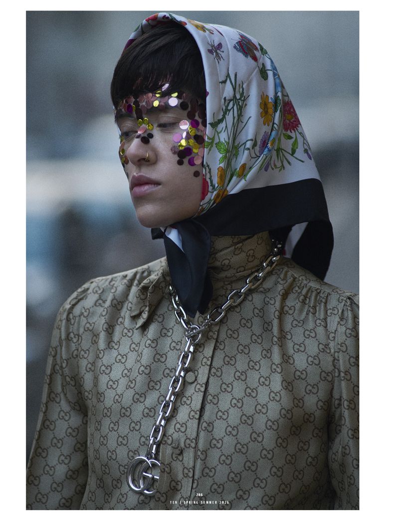 Woman poses in a Gucci patterned blouse and floral headscarf with glittering makeup, against a blurred background in a medium shot.