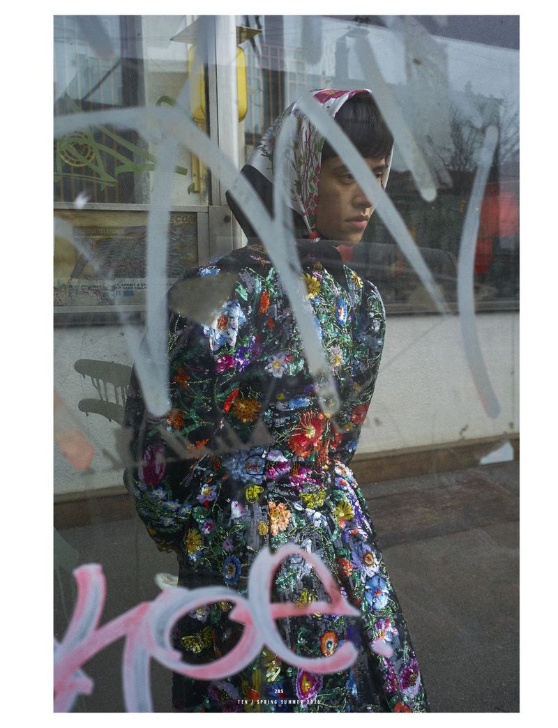 Woman in floral coat and headscarf poses behind graffiti-covered glass, eyes downcast, in an outdoor setting with muted tones.