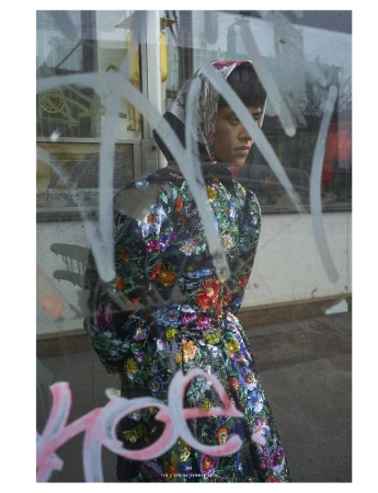 Woman in floral coat and headscarf poses behind graffiti-covered glass, eyes downcast, in an outdoor setting with muted tones.