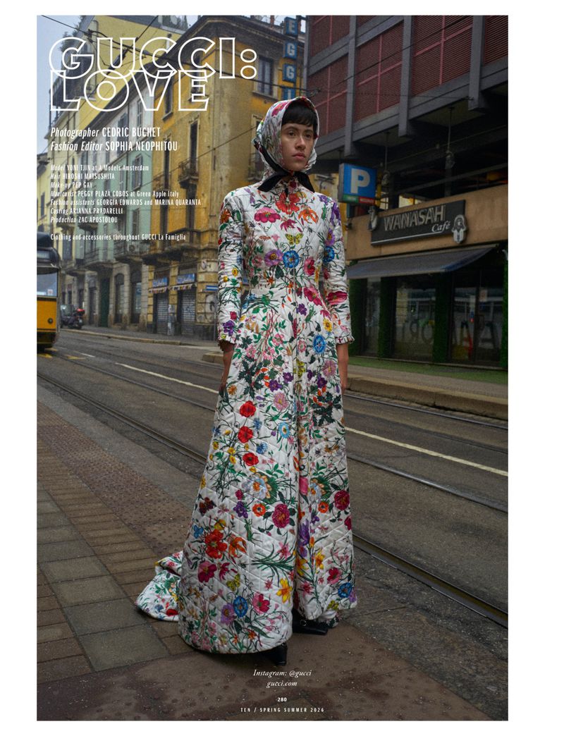 Fashion editorial image featuring a model in a floral gown, posing on a city street near a tram line in front of urban buildings.