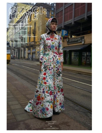 Fashion editorial image featuring a model in a floral gown, posing on a city street near a tram line in front of urban buildings.