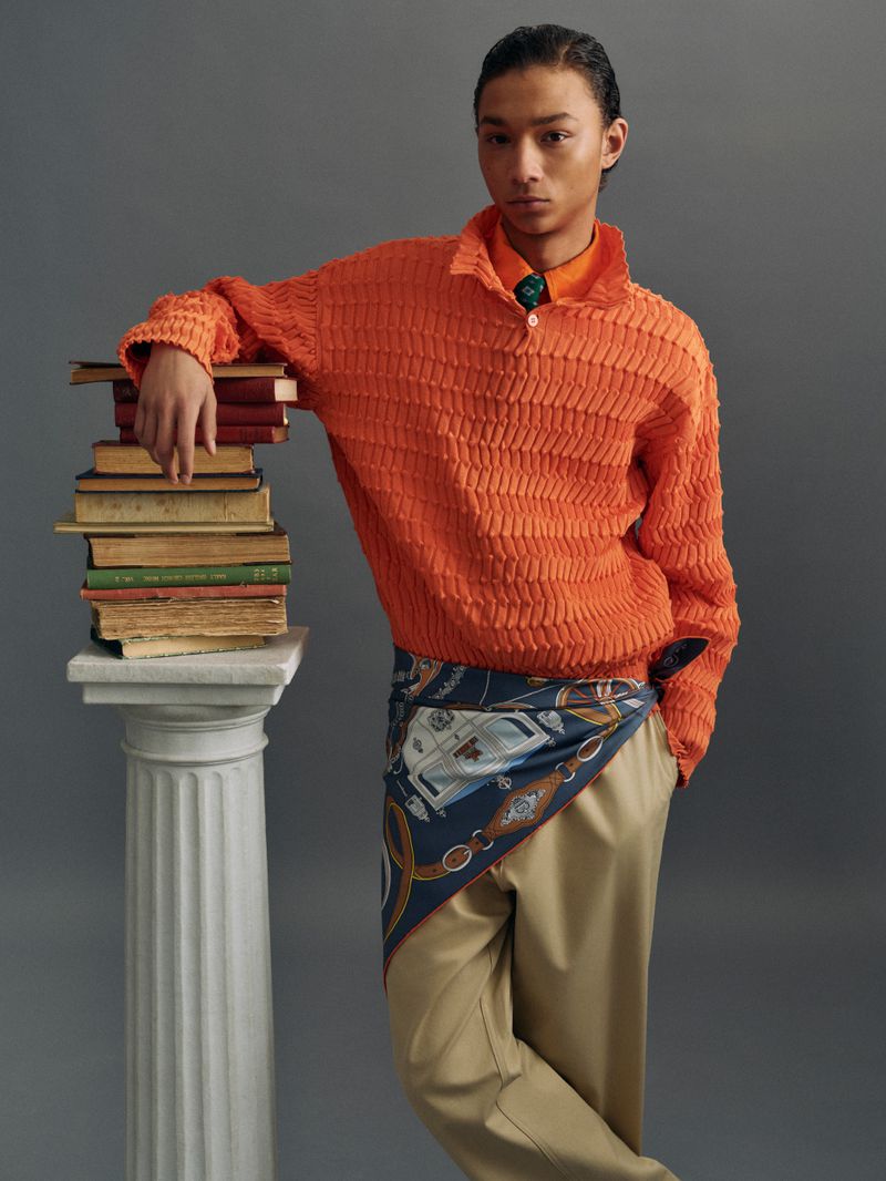 Young Asian man poses in an orange sweater with a stack of books on a column, wearing khaki pants and a patterned scarf on gray backdrop.