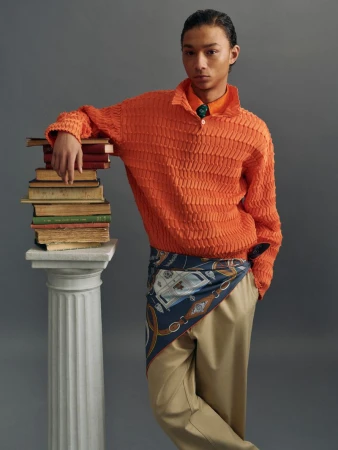 Young Asian man poses in an orange sweater with a stack of books on a column, wearing khaki pants and a patterned scarf on gray backdrop.