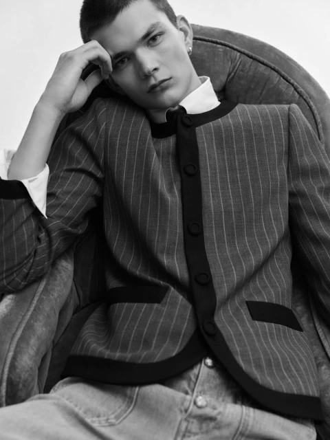 Portrait of a young adult man with dark hair, wearing a pinstripe jacket, collared shirt, and jeans, seated in a plush chair against a white background.