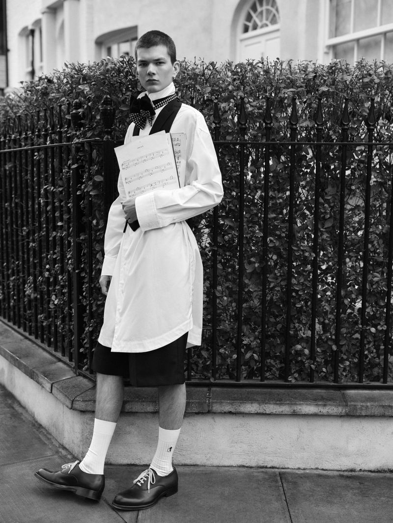 Monochrome portrait of a young man in an oversized white shirt, black shorts, and dress shoes, holding sheet music outdoors.