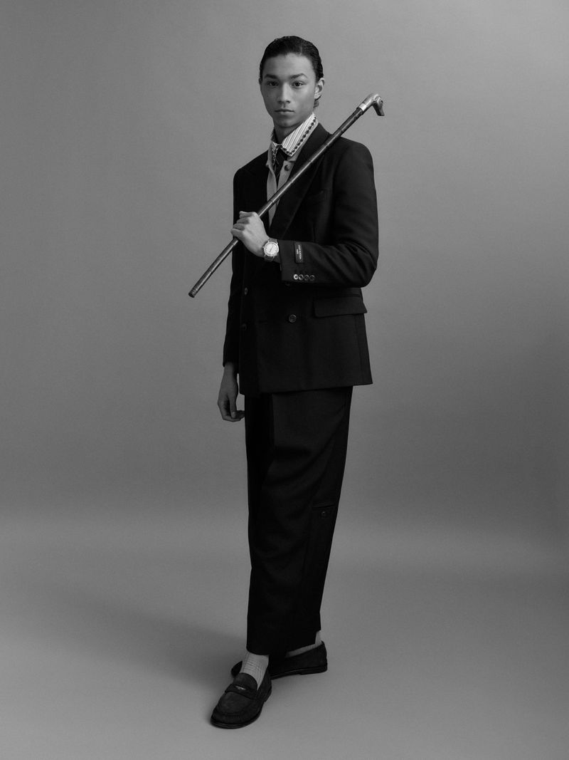 Monochrome portrait of an androgynous person in a dark suit and holding a cane, posing in front of a plain light-grey background.