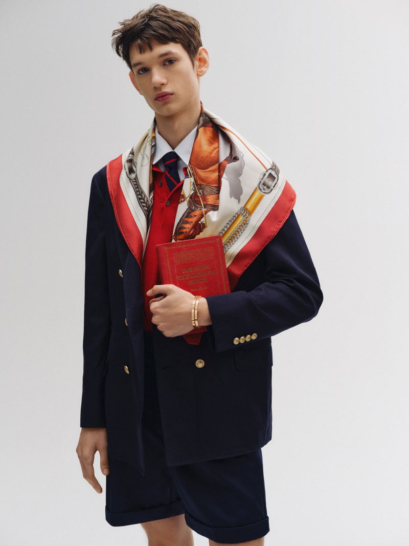 Male fashion model in navy blazer and shorts, holding red book, wearing patterned scarf, standing in a bright photo studio.
