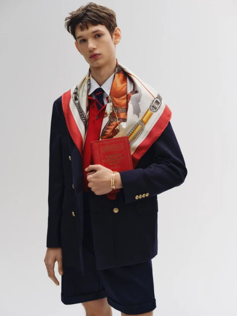 Male fashion model in navy blazer and shorts, holding red book, wearing patterned scarf, standing in a bright photo studio.