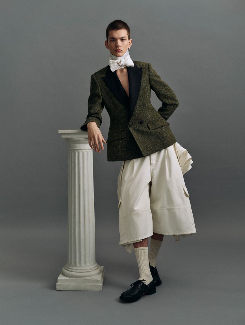 A young man in a green tweed blazer, ivory shorts, white bow tie, and black dress shoes poses next to a white pillar on a grey background.