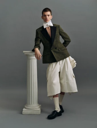 A young man in a green tweed blazer, ivory shorts, white bow tie, and black dress shoes poses next to a white pillar on a grey background.