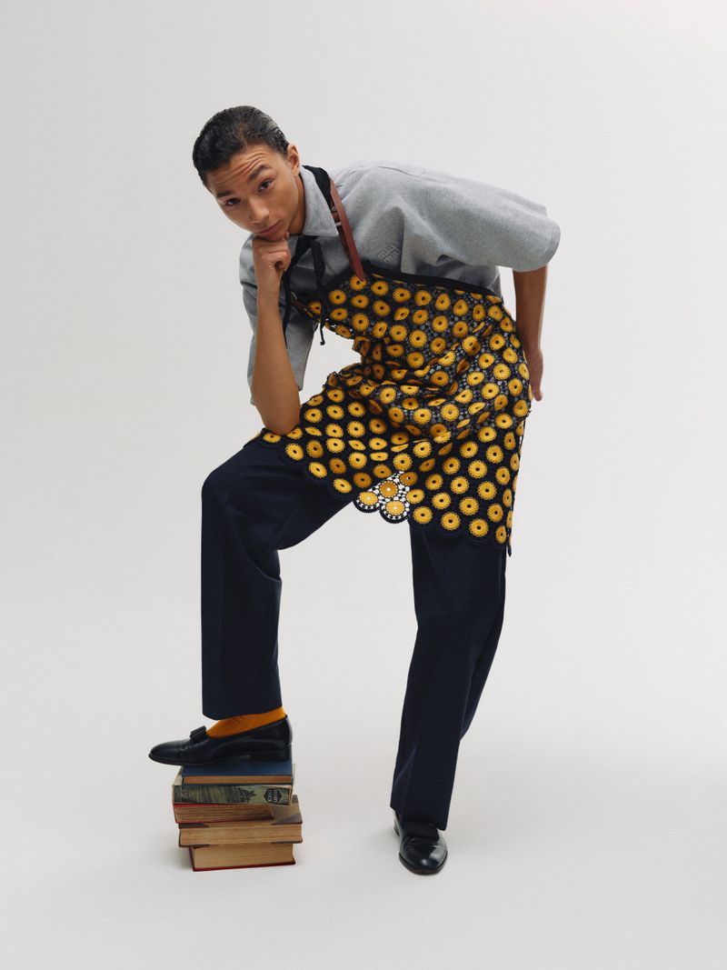 Man in patterned apron posing with hand on chin, one foot on stacked books, wearing yellow socks, in a studio shot.