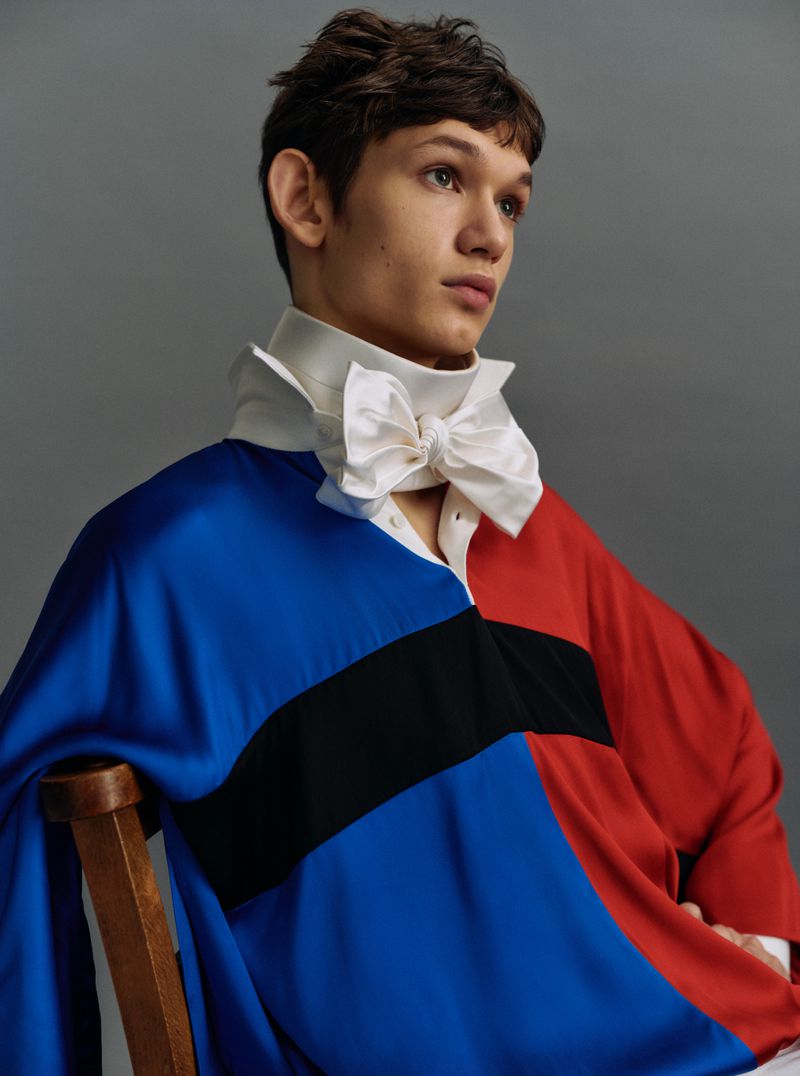 Portrait of a young adult with a color block satin shirt and white bow, sitting on a wooden chair against a gray backdrop.