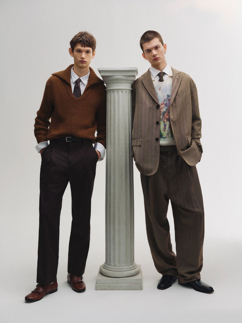 Two young male models pose indoors in complementary earth-tone outfits. One leans against a fluted column in a fashion studio.