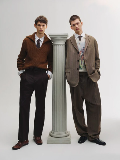 Two young male models pose indoors in complementary earth-tone outfits. One leans against a fluted column in a fashion studio.