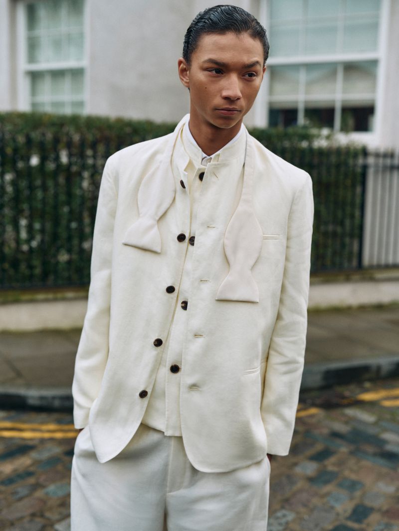 An adult male model poses outdoors in a white three-piece suit, including a jacket, vest, bow tie, and trousers, against a building backdrop.