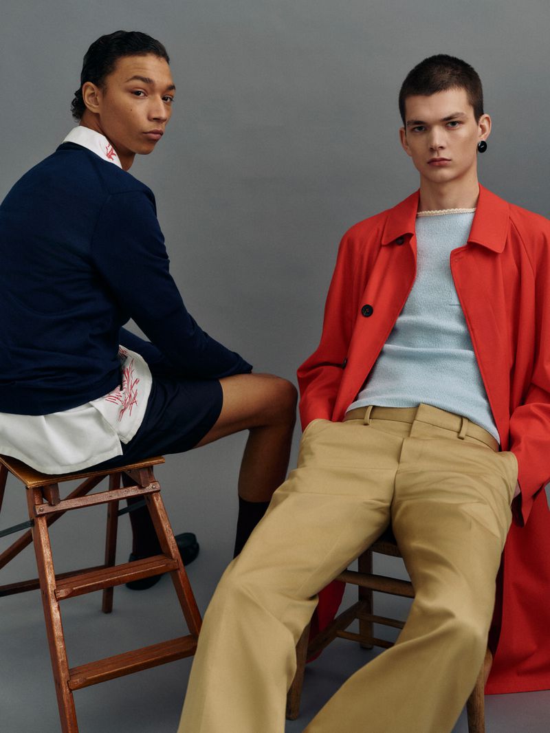 Two young adult men pose indoors in a fashion shot, sitting on wooden chairs and wearing a sweater, shorts, slacks, and a red jacket against a grey backdrop.