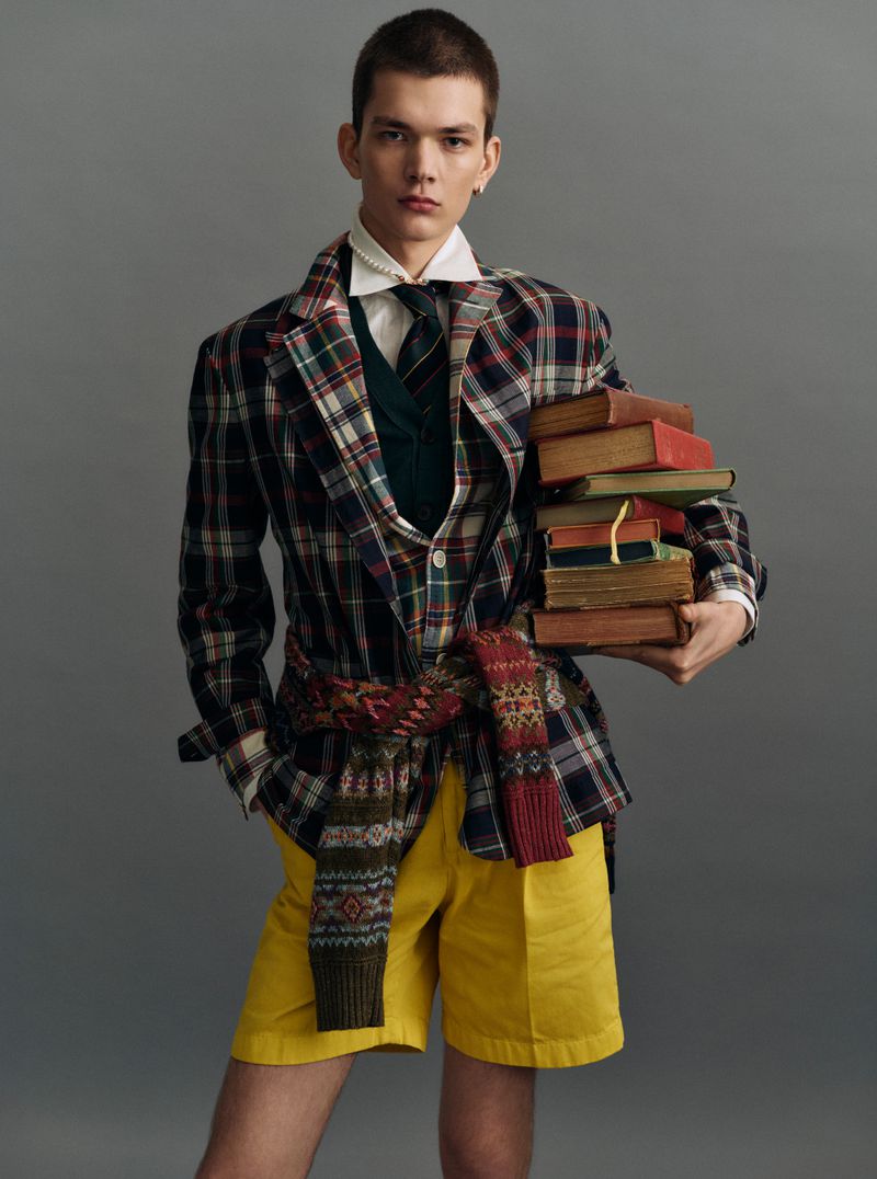 Young male model in a plaid blazer and yellow shorts holding a stack of books against a grey background.