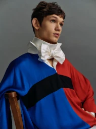 Young male model wearing a color-blocked top and a white bow collar seated against a grey background