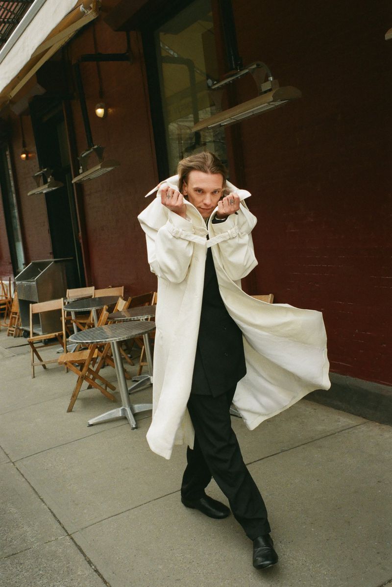An adult male stands on a city sidewalk in a long white coat with the collar up, black shirt and pants, and black shoes.