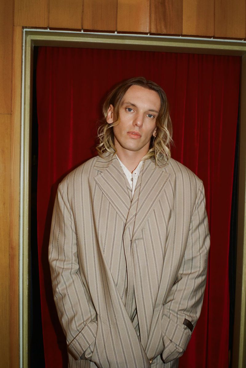 Man in striped suit posing indoors against a red curtain and wood paneling, showcasing fashion and personal style.