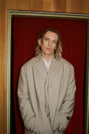 Man in striped suit posing indoors against a red curtain and wood paneling, showcasing fashion and personal style.