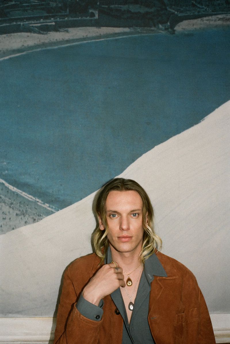 Portrait of Jamie Campbell Bower indoors against artwork, wearing a brown jacket, grey collared shirt, and pendant necklaces. 