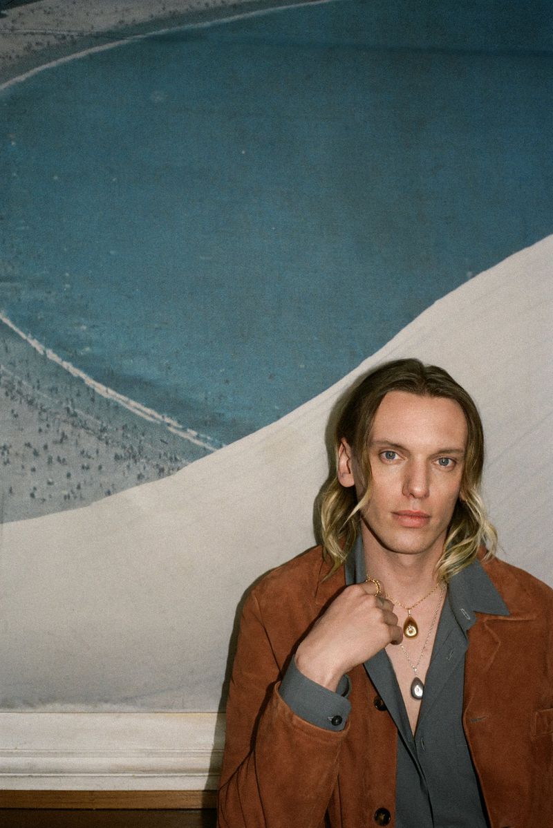 Portrait of adult male with blonde hair wearing a brown jacket and silver necklaces against a muted blue and white backdrop.