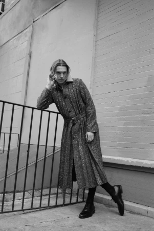 A model leans against a railing, wearing a long coat and black boots, posing in an outdoor urban environment in a monochrome shot.