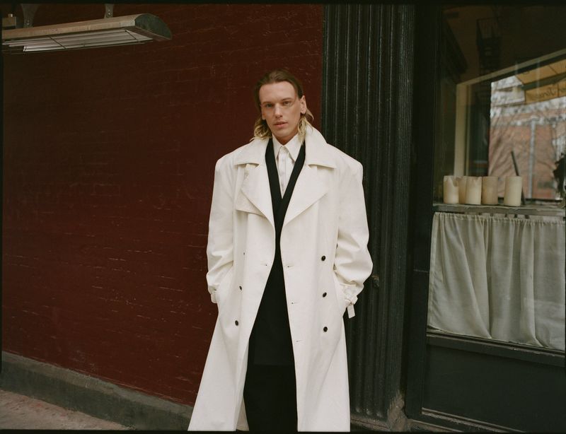Adult male poses in an outdoor urban environment, wearing a white trench coat, a black vest and black pants.  