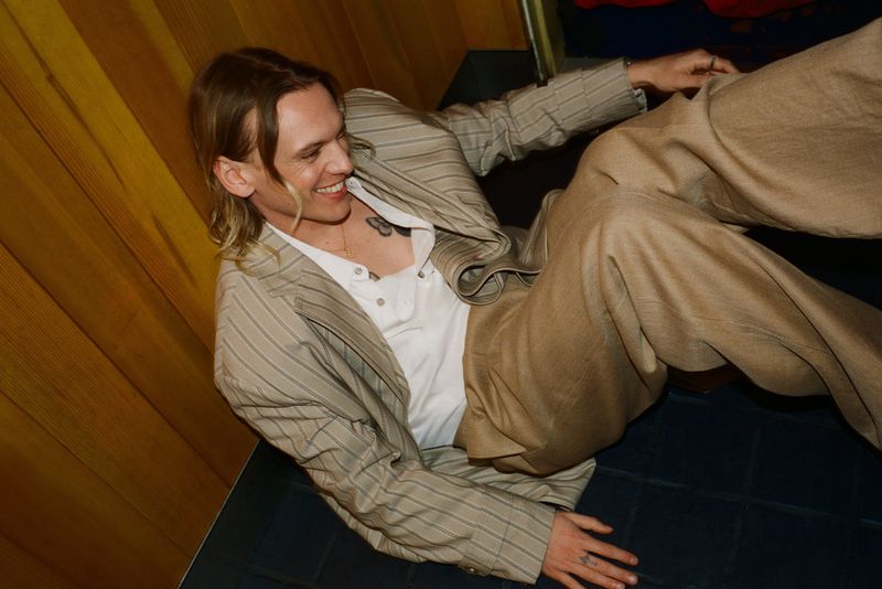 Smiling man in a striped blazer and tan pants reclines against a yellow wood-paneled wall, looking off to the side with a cheerful expression.
