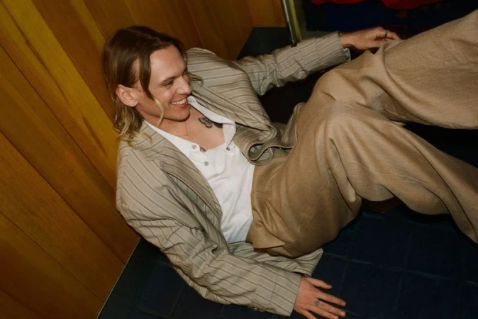 Smiling man in a striped blazer and tan pants reclines against a yellow wood-paneled wall, looking off to the side with a cheerful expression.