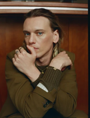 Portrait of adult male with long brown hair, wearing a brown jacket, jewelry, and a brown ribbed sweater with white cuffs while indoors.