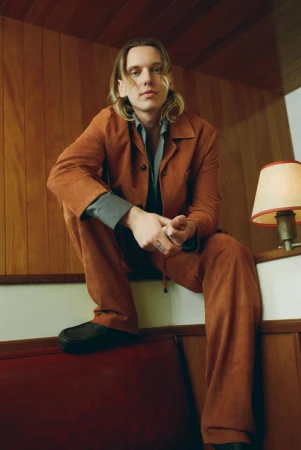 Portrait of a young adult man with blond hair, wearing a brown corduroy suit, sitting on a red surface in a cozy indoor setting.