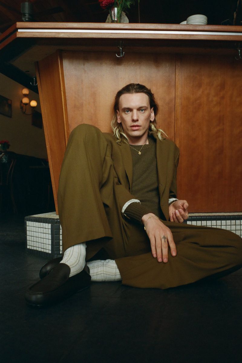 A male model with blonde hair poses indoors wearing an olive green suit, matching top, and dark shoes while seated on the floor.