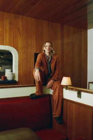 A blonde man in a rust-colored suit sits on a red bench in a retro-styled diner, with wood paneling and dishes visible in the background.